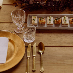 Blick auf goldenen Teller und Besteck mit Gläsern und Fingerfood, rustikaler Tisch im Gartensaal